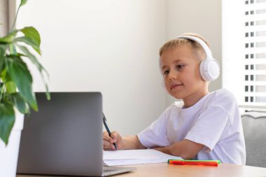 Sevimli bir çocuk çevrimiçi bir okulda okuyor. Çocuk bir dizüstü bilgisayarın önünde kulaklıkla oturuyor ve öğretmeni dinliyor. Eğitim kavramı.