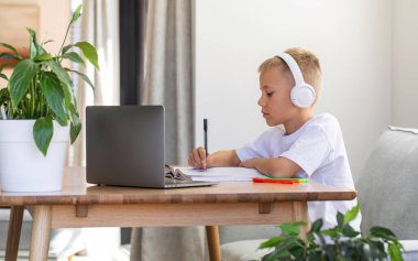 Sevimli bir çocuk çevrimiçi bir okulda okuyor. Çocuk bir dizüstü bilgisayarın önünde kulaklıkla oturuyor ve öğretmeni dinliyor. Eğitim kavramı.