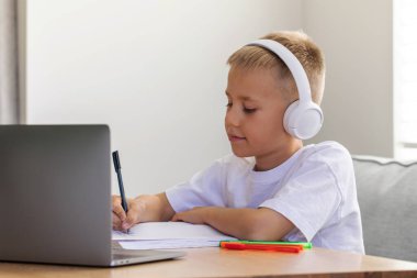 Sevimli bir çocuk çevrimiçi bir okulda okuyor. Çocuk bir dizüstü bilgisayarın önünde kulaklıkla oturuyor ve öğretmeni dinliyor. Eğitim kavramı.