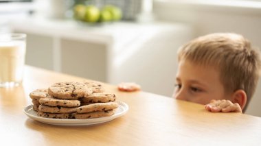 A boy is peeping at an appetizing stack of freshly baked chocolate cookies on the table. The boy hides under the table to secretly eat cookies. clipart