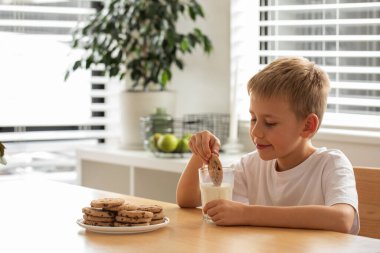 Çocuk ev yapımı kurabiyeyi bir bardak süte batırırken gülümsüyor, atıştırmanın tadını çıkarıyor. Süt ürünü