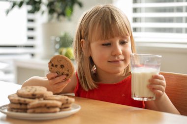 Kurabiye ve sütle neşeli bir kız, evde tatlı bir atıştırmalığın tadını çıkarıyor. Süt ürünü