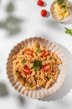 Beyaz bir masada taze otlarla süslenmiş beyaz peynirli ve kiraz domatesli bir tabak spiral makarna. İtalyan yemeği.