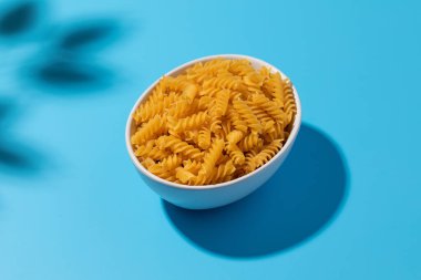 Pişmemiş fusilli makarna mavi arka planda beyaz bir kasede kıvrılır. Gölgeler ve yemek kavramları için idealdir..