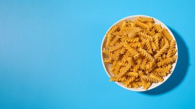 Pişmemiş fusilli makarna mavi arka planda beyaz bir kasede döner. Mutfak ve yemek kavramları için mükemmeldir. Boşluğu kopyala