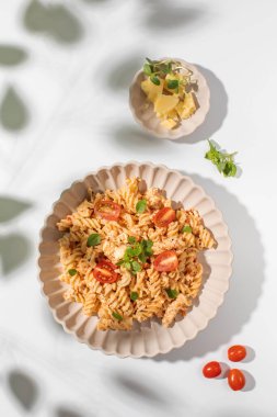 Beyaz bir masada taze otlarla süslenmiş beyaz peynirli ve kiraz domatesli bir tabak spiral makarna. İtalyan yemeği.