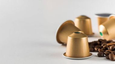 Beyaz arka planda altın kahve kapsülleri ve dağınık fasulyeler modern ve aromatik kahve demleme deneyimini vurguluyor. Boşluğu kopyala