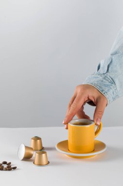 Beyaz arka planda altın kahve kapsülleri ve fasulyeli sarı kahve fincanına uzanan el modern ve uygun bir kahve deneyimini vurguluyor.