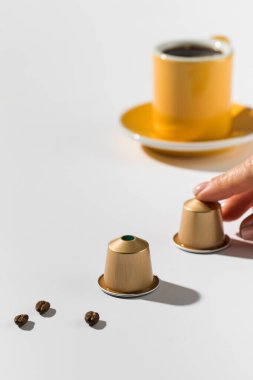 Altın kahve kapsülleri ve beyaz arka planda bir fincan kahve. Kapsül seçen bir el modern bir demleme deneyimini ön plana çıkarıyor.