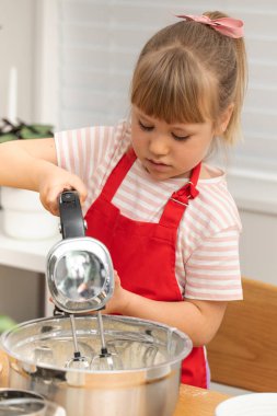 Önlüklü sarışın kız krep hamurunu elektrikli mikserle dövüyor. Mutlu genç ev hanımı evde, mutfakta aile için güzel bir kahvaltı hazırlıyor.