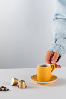 Beyaz arka planda altın kahve kapsülleri ve fasulyeli sarı kahve fincanına uzanan el modern ve uygun bir kahve deneyimini vurguluyor.