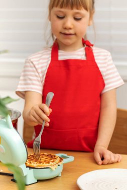 Odaklanmış liseli kız şef waffle 'ı çatalla mini elektrikli fırında kontrol ediyor. Mutlu kız neticeye bakar. Tüm aileyi lezzetli bir kahvaltıyla doyurmaya hazır.