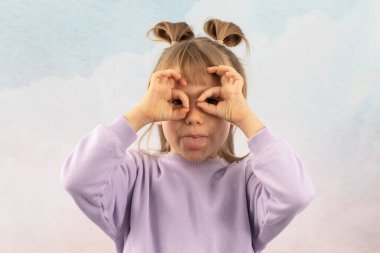 Sarışın kız, göz çevresindeki yüzük şekilli parmaklarıyla gözlük taklidi yaparak yüzünü buruşturuyor. Neşeli liseli kız beyaz arka planda dolanan dille oynuyor.