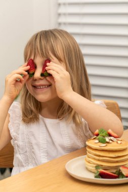 Gülümseyen küçük kız, masada oturmuş gözlerinde krep olan çileklerle gözlerini kapatıyor. Neşeli çocuk mutfakta lezzetli yemeklerle eğleniyor. Çocuklar için beslenme