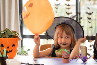 Şapkalı genç bayan elinde balonla cadıcılık oynuyor. Kız, Cadılar Bayramı 'nda arkadaşlarıyla ev partisi hazırlığında daire dekore ediyor.