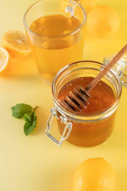 Sarı arka planda ballı bir kavanoz bal, yanında da nane yaprakları ve limon dilimleri..