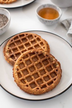 Beyaz bir tabakta ev yapımı yulaf ezmesi ve chia tohumu waffle, yanında bal ve malzemeler, sağlıklı ve besleyici bir kahvaltı seçeneği sunuyor.