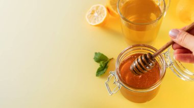 Sarı arka planda ballı bir kavanoz bal, yanında da nane yaprakları ve limon dilimleri. Boşluğu kopyala