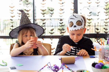 Cadılar Bayramı kostümlü şirin erkek ve kız kardeşler masada şenlikli el sanatları yapıyorlar. Cadılar Bayramı el sanatları üzerine bir master sınıfı. Mutlu Cadılar Bayramı.