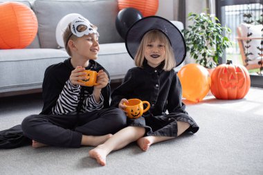 Kız ve erkek öğrenciler ellerinde balkabağı şeklinde bardaklarla yerde otururlar. Korkunç kıyafetler giymiş çıplak ayaklı çocuklar Cadılar Bayramı 'nı kutluyor.