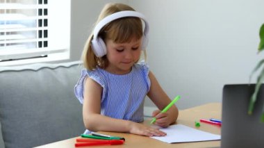 Kablosuz kulaklıklı liseli kız evde video dersi sırasında kağıda resim çiziyor. Güzel küçük kız internet resim dersinde bilgisayarla uzaktan öğreniyor.