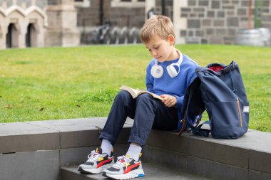 Sırt çantalı ve kulaklıklı küçük çocuk kitap okuyor yemyeşil çimlerin yanında oturuyor. Ortaokul öğrencisi okul bahçesinde ders için ödev yapıyor. Özel okul öğrencisi