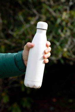Erkek eli, bulanık çalılara yakın çekim için termos tutuyor. Turist, açık havada fotokopi çekilen su şişesini gösteriyor. İçecekler için yeniden kullanılabilir matara