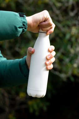 Erkek eli, bulanık çalılara yakın çekim için termos tutuyor. Turist, açık havada fotokopi çekilen su şişesini gösteriyor. İçecekler için yeniden kullanılabilir matara