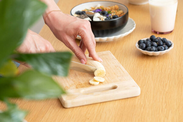 Женщина режет банан на здоровый завтрак с мюсли и греческим йогуртом. Концепция здорового питания.