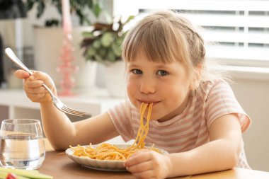 Küçük mutlu kız mutfak masasında oturmuş lezzetli spagetti yiyor. Komik çocuk, güneşli odada lezzetli ev yapımı makarnayla yemek yiyor. Çocuklar için sağlıklı beslenme