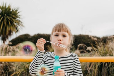 Şirin küçük kız parkta vakit geçirirken sabun köpüğü üflüyor. Sevimli çocuk oyun sahasında renkli köpüklerle oynarken eğleniyor. Mutlu çocukluk