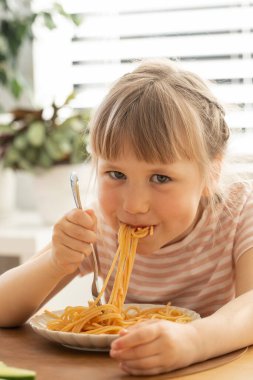 Küçük mutlu kız mutfak masasında oturmuş lezzetli spagetti yiyor. Komik çocuk, güneşli odada lezzetli ev yapımı makarnayla yemek yiyor. Çocuklar için sağlıklı beslenme