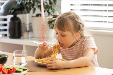 Küçük mutlu kız mutfak masasında oturmuş lezzetli spagetti yiyor. Komik çocuk, güneşli odada lezzetli ev yapımı makarnayla yemek yiyor. Çocuklar için sağlıklı beslenme
