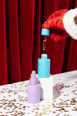 Santa Claus hand holds pipette with liquid over mockup bottle on glitter podium. Christmas gift cosmetic products packaging with design template clipart