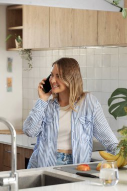 Gülümseyen kadın mutfakta akıllı telefondan konuşuyor. Güzel bir kadın evde sağlıklı atıştırmalıklar hazırlamak için telefonla konuşuyor. Dikey çekim