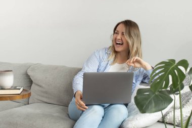 Genç bir kadın oturma odasındaki yumuşak kanepede dizüstü bilgisayar tutarak gülüyor. Sarışın, serbest çalışan bir kadın. İnternette çalışmaktan mutlu. Ofis konforu