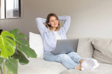 Dizüstü bilgisayarı olan mutlu düşünceli kadın oturma odasındaki yumuşak kanepeye oturur. Rahatlamış dişi evde bilgisayarla internette gezinerek zaman geçirir. Ev hayatı.