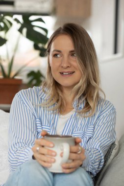 Güzel sarışın kadın rahat koltukta çay fincanı tutuyor. Düşünceli bayan evde kalıp dinlenmeyi ve sakinliği sever. Konforlu ev ortamı