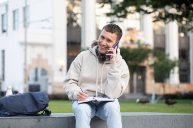Dışarıda kulaklıkla ders çalışırken telefonla konuşan genç bir adam. Şehir Parkı