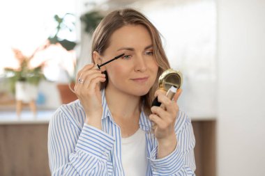 Olumlu, mutlu bir kadın evdeki küçük aynaya bakarak kirpiklerine rimel sürüyor. Genç sarışın kadın makyajı profesyonel kozmetiklerle ayarlıyor.