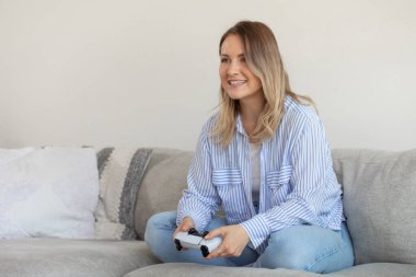 Heyecanlı genç bir kadın oturma odasında kontrolörle oyun oynuyor. Mutlu kadın koltukta otururken dijital eğlenceyle eğleniyor. Kadın oyuncu portresi