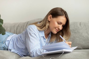 Gülümseyen kadın günlüğe rahat kanepeye uzanan notlar yazıyor. Sarışın kadın oturma odasında günlük planlama için liste yapıyor. Yaşam tarzı organizasyonu