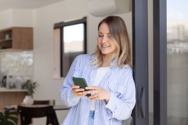 Gülümseyen kadın teras girişinde duran akıllı telefondan mesajı okuyor. Güzel bir kadın evdeki cep telefonuyla sosyal medyayı tarıyor. İletişim çevrimiçi