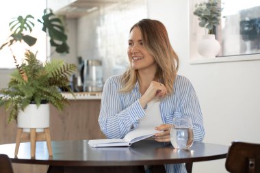 Gülümseyen genç kadın masa başında kalemle notlar yazıyor. Neşeli kadın modern stüdyo dairede fikirler ve planlar yaratıyor.