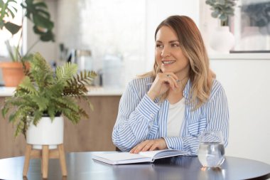 Gülümseyen genç kadın masa başında kalemle notlar yazıyor. Neşeli kadın modern stüdyo dairede fikirler ve planlar yaratıyor.