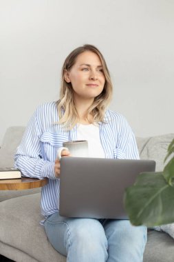 Rahat genç bir kadın rahat oturma odasında dizüstü bilgisayarla ve kahveyle çalışıyor. Laptop kullanılıyor