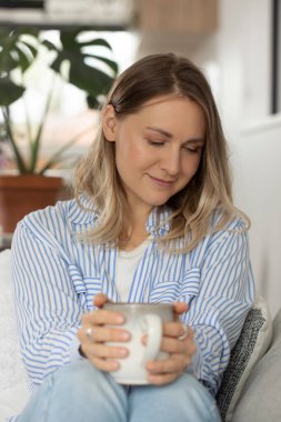 Güzel sarışın kadın rahat koltukta çay fincanı tutuyor. Düşünceli bayan evde kalıp dinlenmeyi ve sakinliği sever. Konforlu ev ortamı