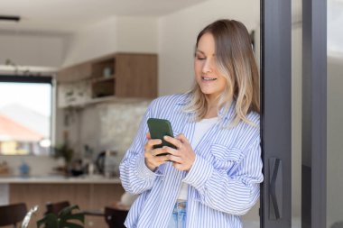 Gülümseyen kadın teras girişinde duran akıllı telefondan mesajı okuyor. Güzel bir kadın evdeki cep telefonuyla sosyal medyayı tarıyor. İletişim çevrimiçi