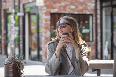 Güneş gözlüklü genç bir kadın kafede açık verandada otururken tek kullanımlık kahve tutuyor. Paket servis, iş kadını, şehir seyahati.