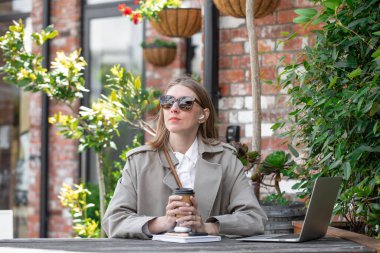 Genç iş kadını, kafedeki açık verandada tek kullanımlık kahve tutuyor. İşte mola. Uzaktan çalışma, serbest konsept.